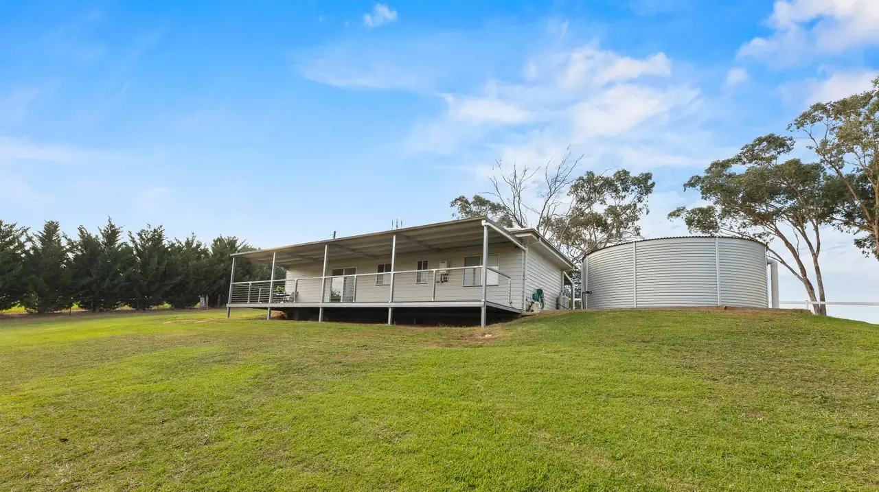 The cottage and rainwater tank from the south, on a wide sloping lawn with a cypress hedge behind